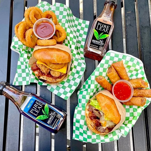 Two Uncle Boy's burgers, onion rings, egg rolls, and Pure Leaf iced teas on a black table.