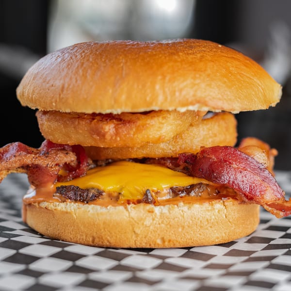 A close-up of Uncle Boy's burger with bacon, melted cheese, and onion rings on a checkered paper.